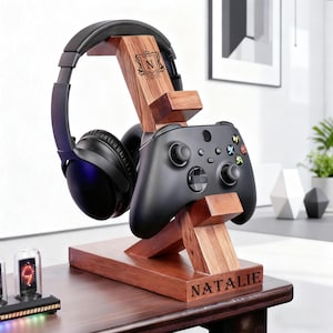 May include: A wooden gaming headset and controller stand. The stand is made of dark wood and features a headset, a black controller, and the name "NATALIE" engraved on the base. The stand also has a monogram with the letter "N".