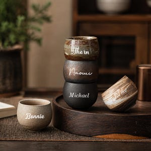 May include: A set of five small ceramic cups with rounded shapes and varied earth-toned finishes. Each cup features a white script name printed on the front: Thorn, Naomi, Michael, Mike, and Bonnie. The cups are arranged in a stack and individually.