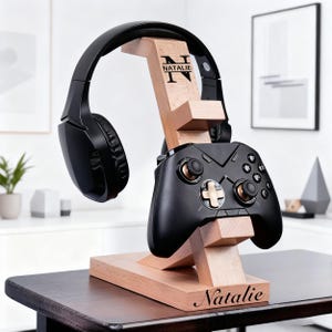 May include: A wooden headphone and game controller stand with the name "Natalie" engraved on the base. The stand holds a black headset and a black game controller. The stand is made of light-colored wood.
