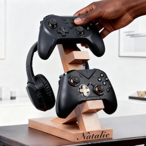 Peut inclure: Un support en bois pour manettes de jeu et casque audio. Il présente deux manettes noires et un casque noir. Le nom "Natalie" est gravé sur la base en bois clair.