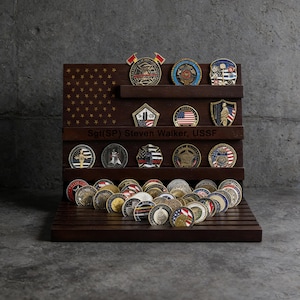 Puede incluir: Un expositor de madera marrón oscuro que contiene numerosas monedas de colección. El expositor presenta un diseño de bandera estadounidense tallada y el texto "Sgt(SP) Steven Walker, USSF". Varias monedas se exhiben en estantes y en la base.
