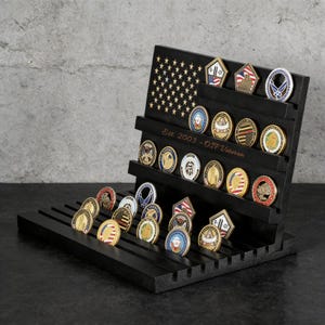 Op de afbeelding: Een zwarte houten muntenstandaard met meerdere rijen, met een ontwerp van de Amerikaanse vlag. De standaard bevat verschillende militaire uitdagingsmunten met kleurrijke ontwerpen. Tekst op de standaard luidt "Est. 2003 - OIF Veteran."