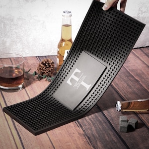 May include: A black bar mat with a textured surface and a rectangular area with the name "Thomas" and the letter "T" printed on it. A glass of amber liquid, a beer bottle, and whiskey stones are also visible.