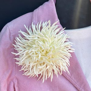 May include: A large, off-white, spiky flower brooch pinned to a light pink, corduroy-textured blazer. The flower features numerous thin, pointed petals radiating from the center, creating a dense, spherical, and textured floral appearance.