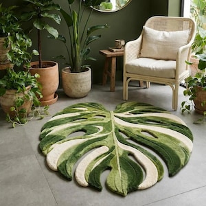 May include: A large, tufted area rug shaped like a monstera leaf, featuring a layered design in shades of dark green, light green, and cream. The rug has deep cutouts and a textured surface that mimics the natural veins and fenestrations of a leaf.