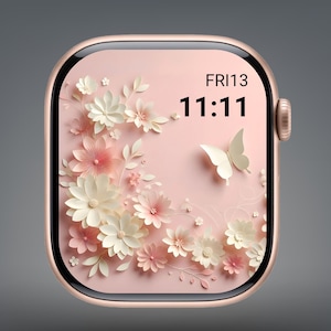 Pode incluir: Um smartwatch em ouro rosa com um design floral na tela. A tela exibe um fundo rosa claro com flores de papel brancas e rosas e uma borboleta branca. A hora é 11:11 e a data é sexta-feira 13.