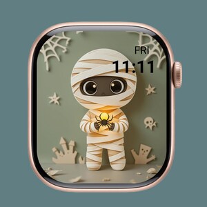 Puede incluir: Un Apple Watch de oro rosa muestra una esfera con temática de Halloween. La esfera presenta un personaje de momia sosteniendo una araña brillante, con la hora 11:11. El fondo incluye telarañas, estrellas y otros elementos espeluznantes.