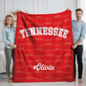 Puede incluir: Una manta de forro polar de color rojo brillante con un patrón repetitivo de la palabra TENNESSEE en un sutil estampado tono sobre tono. La manta muestra la palabra TENNESSEE en letras mayúsculas blancas y el nombre Olivia en cursiva.
