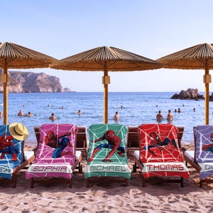 May include: Five beach towels draped over wooden lounge chairs, each featuring a Spider-Man graphic with a web pattern and a personalised name at the bottom: Oliver, Sophia, Michael, Thomas, and Olivia. A straw hat rests on the first towel.