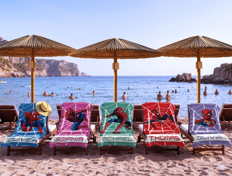 May include: Five beach towels draped over wooden lounge chairs, each featuring a Spider-Man graphic with a web pattern and a personalised name at the bottom: Oliver, Sophia, Michael, Thomas, and Olivia. A straw hat rests on the first towel.