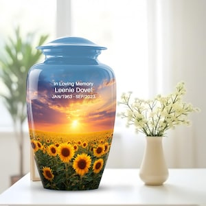 May include: A blue cremation urn with a sunflower field and sunset design. The urn has the text "In Loving Memory Leenie Dovel JAN/1963 - SEP/2023". The urn is sitting on a white table next to a vase of white flowers.