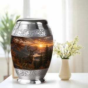 May include: A metallic cremation urn featuring a polished silver-tone lid and base with intricate engraved scrollwork. The central body displays a detailed landscape scene of a deer standing in a forest at sunset with mountains and a lake.