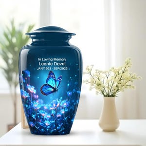 May include: A dark blue cremation urn with a glossy finish. The urn features a vibrant design of a blue butterfly and blue flowers. Text on the urn reads "In Loving Memory Leenie Dovel JAN/1963 - SEP/2023". A white vase with flowers is to the right.