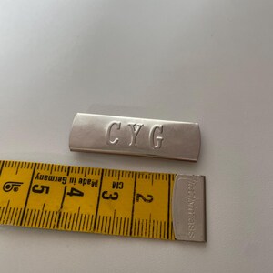 Könnte beinhalten: Ein silberner Metallstab mit den Buchstaben "CYG" eingraviert. Ein gelbes Maßband liegt neben dem Stab. Der Stab ist etwa 2,5 cm lang.