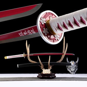 Puede incluir: Una espada katana japonesa con una hoja roja y un mango blanco. La espada presenta un diseño floral en la guarda y se exhibe sobre un soporte de cuerno de ciervo. La hoja tiene caracteres japoneses grabados.