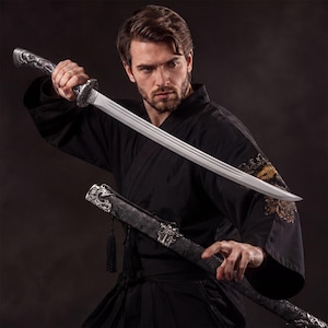 Puede incluir: Una katana con una hoja plateada y un mango ornamentado se sostiene frente a un kimono negro. La vaina, también negra, presenta detalles decorativos plateados. La espada se sostiene en una postura de combate.