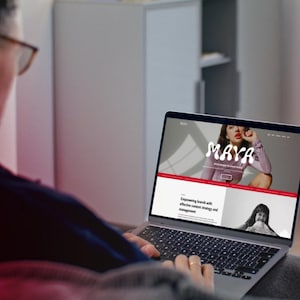 May include: A person using a laptop displaying a website with the name "MAYA" in a stylized font. The website features a photo of a woman and text about content strategy and management. The laptop is silver and has a black keyboard.