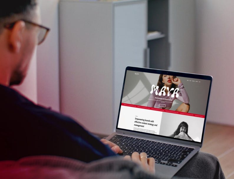 May include: A person using a laptop displaying a website with the name "MAYA" in a stylized font. The website features a photo of a woman and text about content strategy and management. The laptop is silver and has a black keyboard.