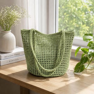 Könnte beinhalten: Eine hellgrüne Häkeltasche mit zwei Henkeln. Die Tasche hat eine rechteckige Form mit einer breiten Öffnung und einem strukturierten, offen gewebten Design. Die Tasche steht auf einer Holzoberfläche in der Nähe eines Fensters.