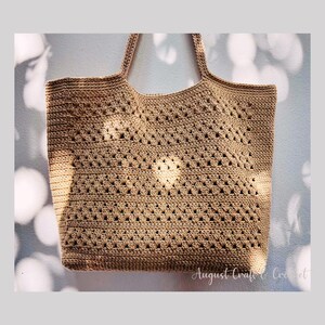 Könnte beinhalten: Beige gehäkelte Tragetasche mit zwei Henkeln. Die Tasche hat ein offenes Design mit einem sich wiederholenden Muster aus Rauten. Der Text "August Craft & Crochet" ist unten rechts sichtbar.