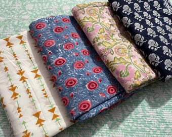 May include: A collection of folded fabric with various patterns. One fabric is white with orange and green arrow designs. Another is blue with red floral patterns. A third is pink with green floral patterns, and the last is navy with white floral patterns.