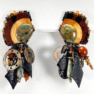 May include: A pair of handcrafted earrings featuring a fan-shaped top with layered brown, tan, and black elements, accented with a green stone. Below, dangling elements include metal rings, black shapes, and various beads.