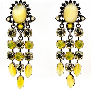 Pode incluir: Um par de brincos pendurados com uma estética vintage. Cada brinco apresenta uma grande pedra oval, amarelo pálido no topo, rodeada por filigrana de metal escuro. Abaixo, três fileiras de pedras quadradas e ovais amarelo-esverdeadas descem.