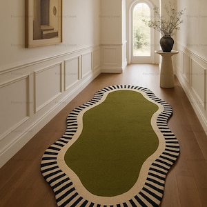 Op de afbeelding: Een olijfgroen, vrijvormig tapijt met een beige rand en een zwart-wit gestreepte rand. Het tapijt ligt op een houten vloer in een gang met witte muren en een ingelijst schilderij.