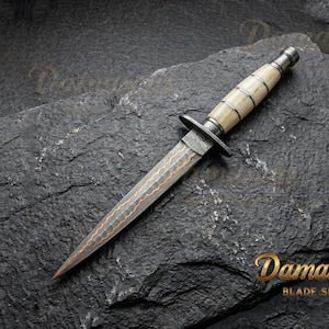 May include: A Damascus blade knife with a bone handle and a metal guard. The blade has a copper-coloured pattern. The knife is displayed on a dark, textured surface. The logo "Damascus Blade Shop" is in the bottom right corner.