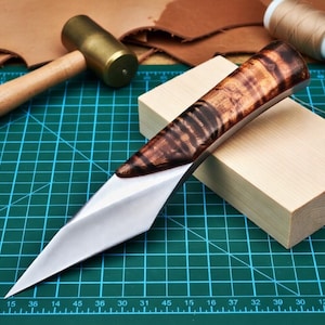May include: A handcrafted knife with a polished steel blade and a wooden handle featuring a unique grain pattern. The knife rests on a green cutting mat, showcasing its sharp, angular design. The handle is approximately 6 inches long.