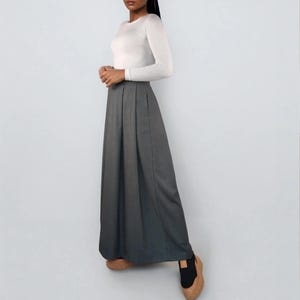 May include: A woman wearing a white long-sleeve shirt and a gray maxi skirt. The skirt is pleated and falls to the ankles. The woman is wearing brown and black shoes. The background is a solid light gray.