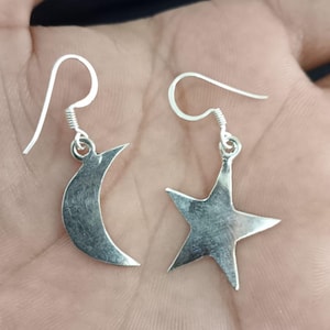 Könnte beinhalten: Ein Paar silberne Ohrringe, einer in Form eines Halbmonds und der andere in Form eines fünfzackigen Sterns. Die Ohrringe haben ein schlichtes, elegantes Design mit Hakenverschluss. Der Schmuck wird vor neutralem Hintergrund präsentiert.