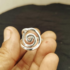Peut inclure: Une bague argentée avec un motif en spirale. La bague présente une spirale ouverte en relief au centre, avec une finition lisse et polie. La bague est tenue dans une main sur un fond sombre.