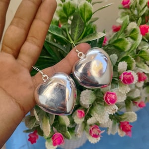 Pode incluir: Um par de brincos prateados em forma de coração. Os brincos são lisos e brilhantes, com um design de coração arredondado e tridimensional. São suspensos por ganchos prateados simples. O fundo apresenta folhas verdes e flores cor-de-rosa.