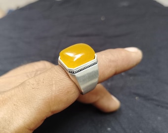 Könnte beinhalten: Ein Silberring mit einem großen, gelben, rechteckigen Edelstein. Der Ring hat ein schlichtes, modernes Design mit poliertem Finish. Der Edelstein ist glatt und hat eine leicht abgerundete Oberseite. Der Ring wird am Finger getragen.