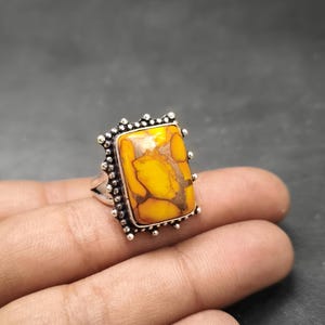 May include: Silver ring with a rectangular, yellow and brown stone. The stone is set in a decorative silver frame with small, round accents. The ring is displayed on a hand.