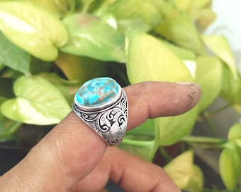 May include: A silver ring with an oval turquoise stone. The ring has an ornate, engraved design on the band. The turquoise stone is a mix of blue and brown hues. The ring is displayed on a finger.
