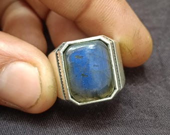 May include: A silver ring with a rectangular, blue gemstone. The gemstone has a shimmering, iridescent quality. The ring's band is thick and has a textured design on the sides. The ring is held by a hand.