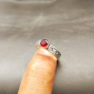 Puede incluir: Un anillo de plata con una piedra preciosa redonda de color rojo intenso. La banda presenta un patrón detallado. El anillo se muestra en un dedo sobre un fondo oscuro. Una joya.
