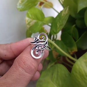 Op de afbeelding: Een zilverkleurige ring met een sierlijk, wervelend ontwerp. De ring heeft een reeks met elkaar verbonden curven en lussen, waardoor een vloeiend, abstract patroon ontstaat. De ring wordt getoond tegen een achtergrond van groen gebladerte.