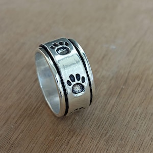 May include: A silver-colored ring featuring a repeating paw print design. The ring has a textured appearance and a rotating central band. The paw prints are black and add a playful touch to the jewelry.