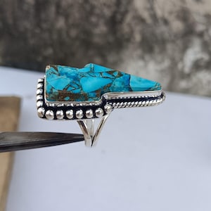 Pode incluir: Anel de prata com uma pedra turquesa retangular com veios castanhos. A pedra é colocada numa moldura de prata decorativa com uma borda de contas e um design de corda torcida. O anel tem uma haste dividida.
