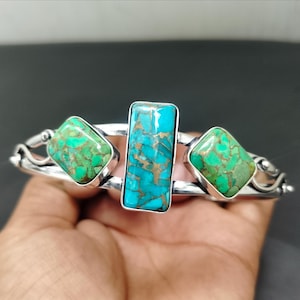 May include: Silver bracelet featuring three turquoise stones. The central stone is rectangular, flanked by two diamond-shaped stones. The turquoise has a vibrant blue-green hue with copper-colored matrix.