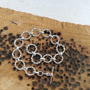 May include: A silver bracelet with a series of interconnected, twisted rope-like rings. The bracelet features a toggle clasp closure. The jewelry is displayed on a textured, wooden surface, showcasing the contrast between the shiny metal and the natural wood.
