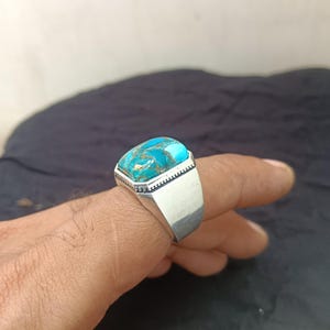 Può includere: Un anello d'argento con una pietra turchese rettangolare. La pietra presenta una miscela di colori blu e oro. L'anello è su un dito, su uno sfondo scuro.