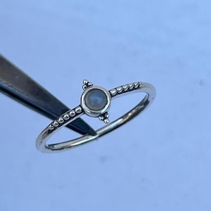 May include: A silver ring featuring a small, round, pale-coloured stone at its centre. The thin ring band has small, evenly spaced bead-like details. The stone is held by three small, raised prongs.