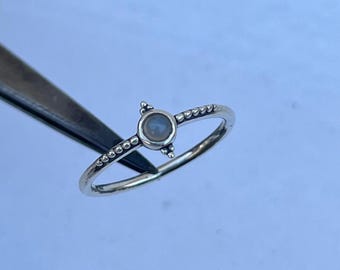 May include: A silver ring with a small, round, light-colored stone at the center. The ring band is thin and features small, evenly spaced bead-like details. The stone is held in place by three small, raised prongs.