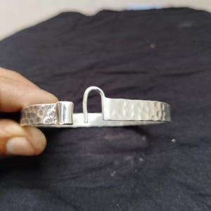 Può includere: Un bracciale a polsino color argento con una texture martellata. Il bracciale ha una chiusura rettangolare e una chiusura a gancio curvo. Il metallo ha una finitura lucida. Il bracciale è mostrato su uno sfondo scuro.