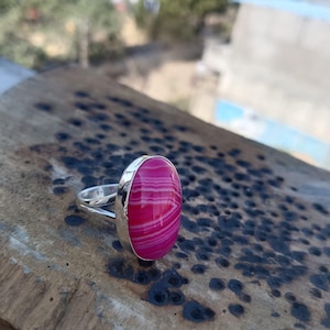 May include: A silver ring with a large, oval pink gemstone with white stripes. The ring is set on a textured wooden surface, with a blurred background of greenery and a building.