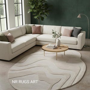 Op de afbeelding: Een moderne woonkamer met een crèmekleurige hoekbank en kussens in groen, roze en grijs. Een ronde houten salontafel staat op een abstract gevormd tapijt met een gelaagd ontwerp. Het tapijt heeft de tekst "NR RUGS ART".